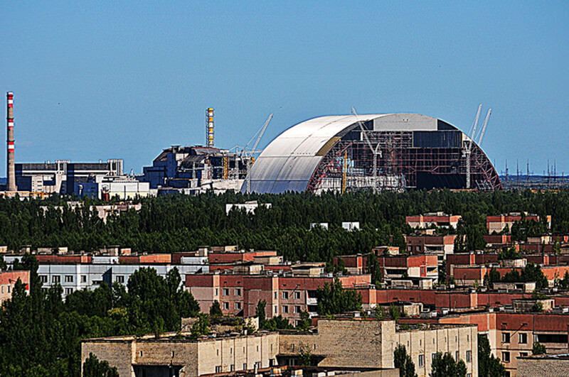 Vista panorámica del Central Nuclear Vladimir Ilich Lenin, mundialmente conocida como Central Nuclear de Chernóbil. En primer plano la ciudad fantasma de Pripyat. Al fondo, al lado del malogrado reactor, el nuevo sarcófago (o, por su nombre en inglés, New Safe Confinement) que pondrá a resguardo el letal contenido por un siglo más. Toneladas de combustible continuarán ardiendo por miles de años.