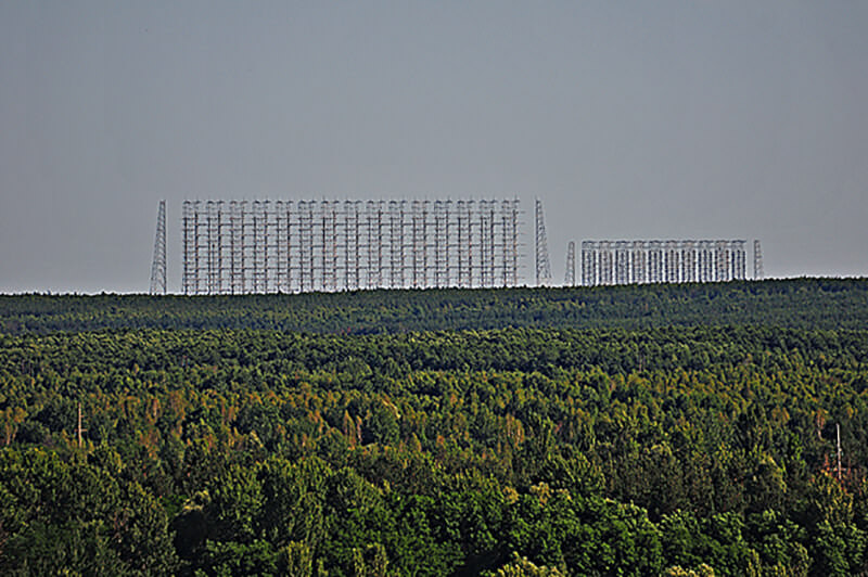 Al fondo la Duga, conocida por los radioaficionados del mundo como Russian Woodpecker (Pájaro carpintero ruso) debido al sonido de su señal. La antena antimisiles más potente de la Guerra Fría. Ubicada en la Zona de Alto Secreto denominada Chernóbil II, la función de esta gigantesca antena era prevenir un ataque nuclear occidental a la Unión Soviética. Una de las teorías señala que la explosión del reactor no fue accidente sino un sabotaje de los servicios secretos occidentales para sacarla de servicio. La planta nuclear de Chernóbil proveía de la enorme energía que requería este poderoso aparato de vigilancia. Su señal era tan potente que causaba interferencias incluso a las comunicaciones en el Pacífico hasta Australia.